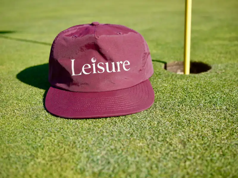 The Rojo Hat in burgundy nylon, Leisure across the front, on the green beside the pin