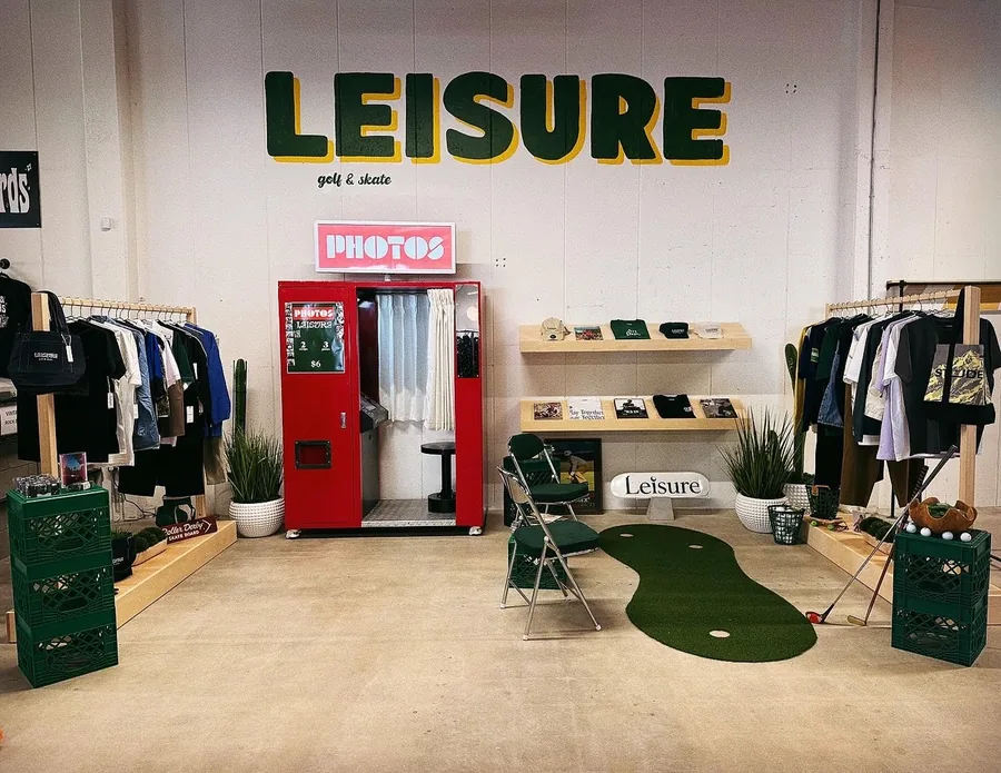 Inside Leisure — the wordmark painted on the back wall, a red photo booth, racks of goods and a putting green on the floor